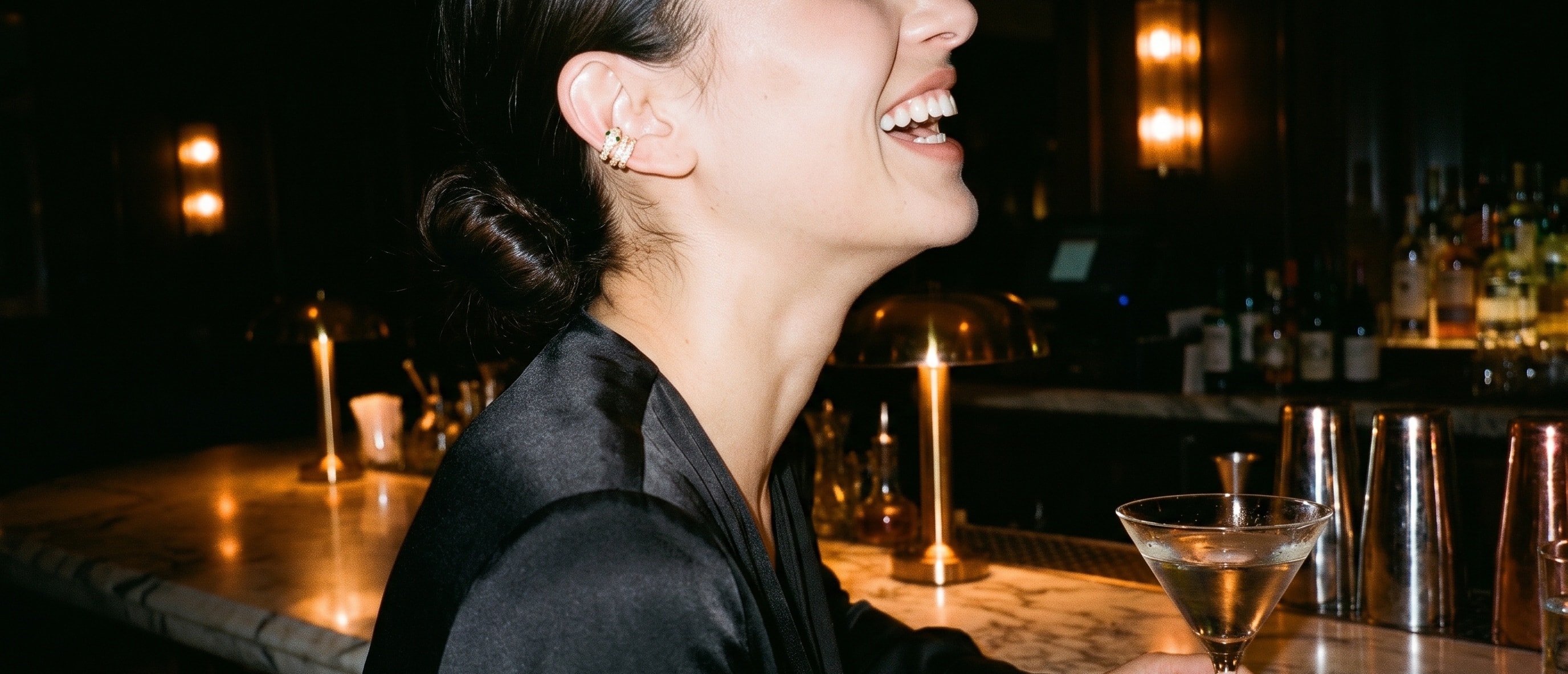 Mulher rindo no balcão de mármore de um bar à noite, com o ear cuff Serpente dourado na orelha