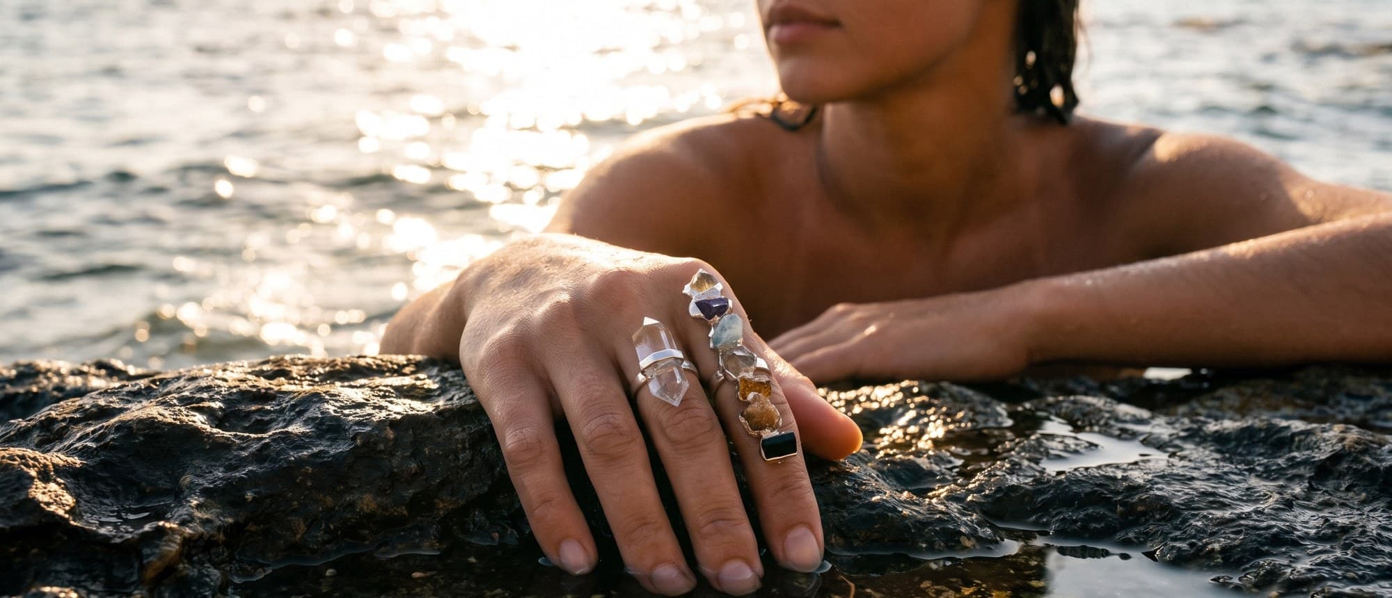Modelo apoiada na pedra de uma piscina natural ao fim da tarde, com o anel 7 Chakras de prata: a barra de pedras no dedo médio e o quartzo paralelo no indicador