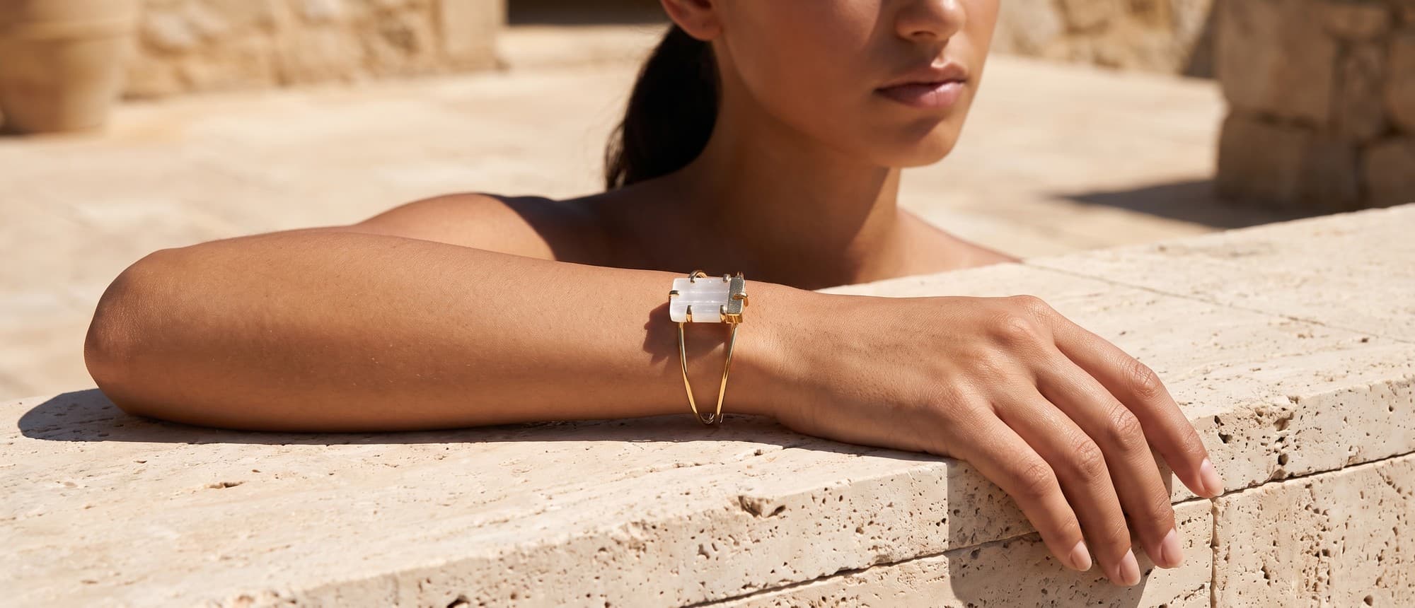 Bracelete dourado com selenita branca no pulso, o braço apoiado em borda de travertino