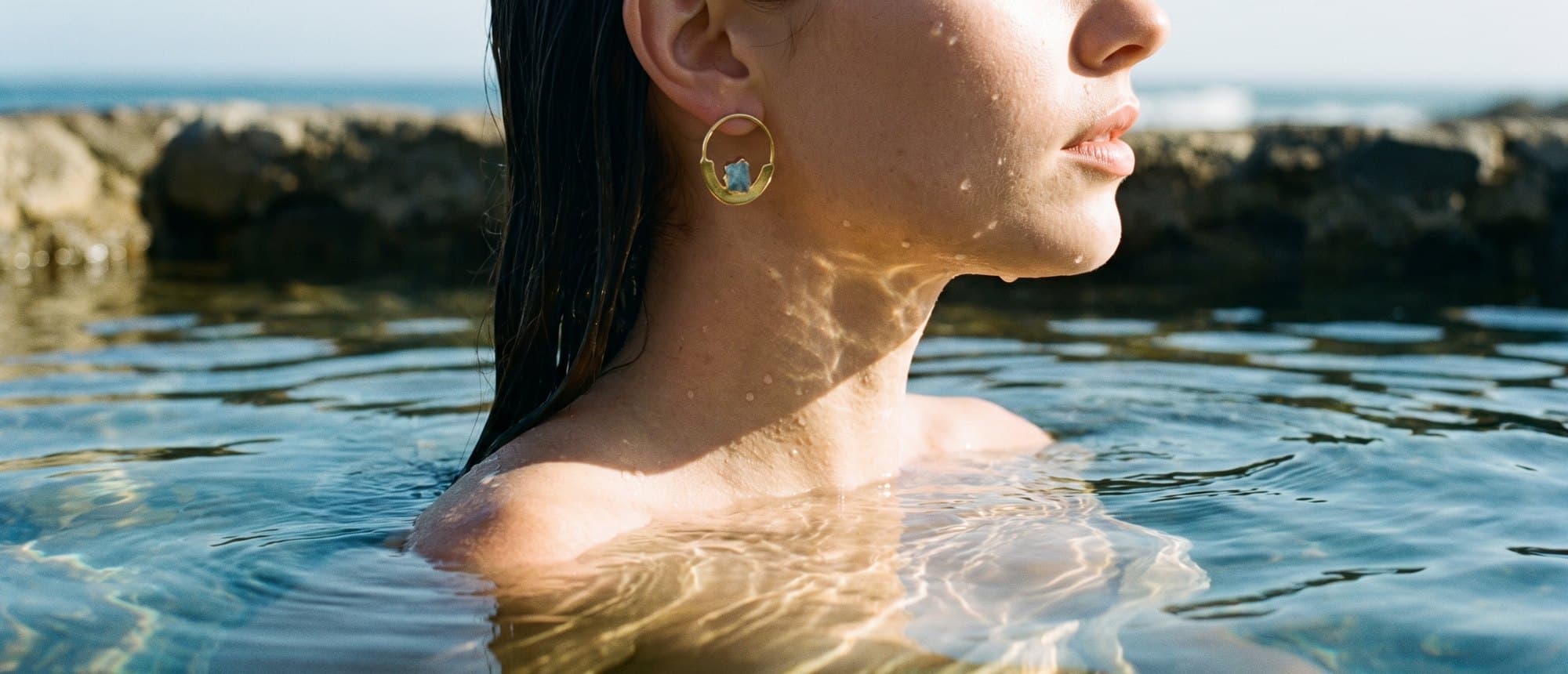 Brinco dourado com água-marinha bruta na orelha, numa piscina natural de pedra